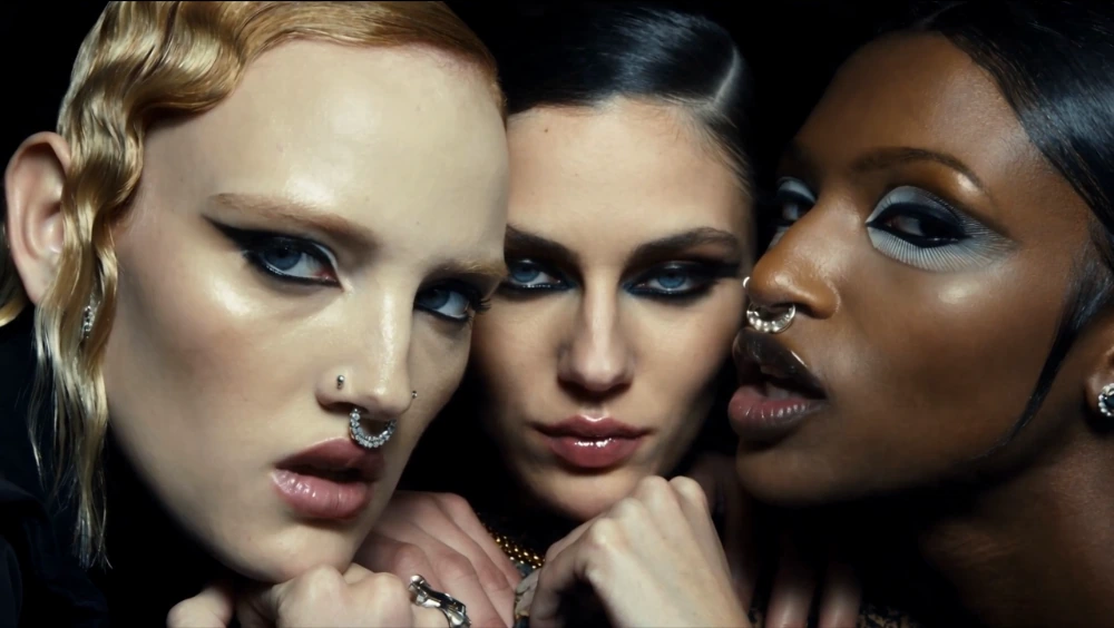 Three women in a close-up fashion video, wearing bright eye makeup and gold jewelry.