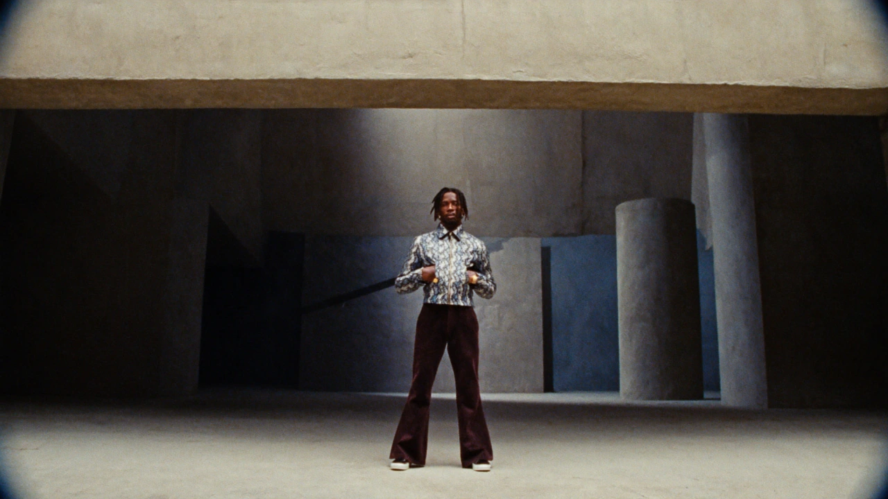 A figure in a patterned shirt and flared trousers stands among monumental concrete columns.