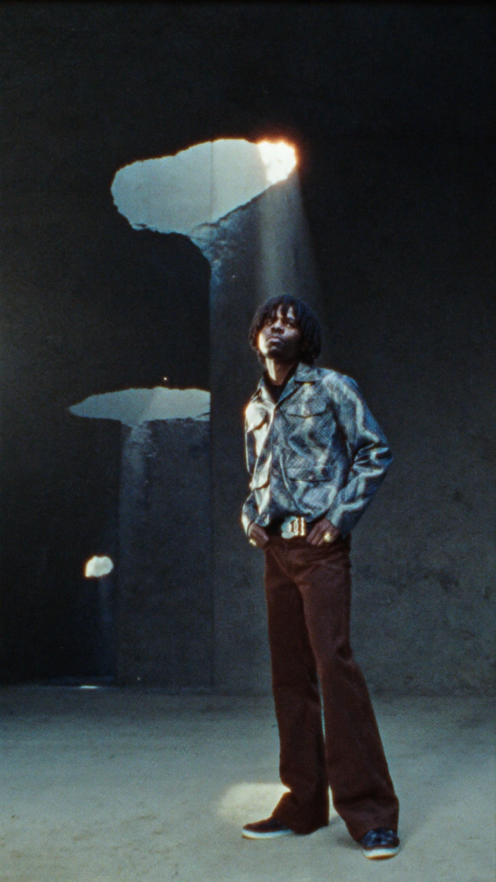 A figure in a patterned shirt stands beneath shafts of light through a concrete ceiling.