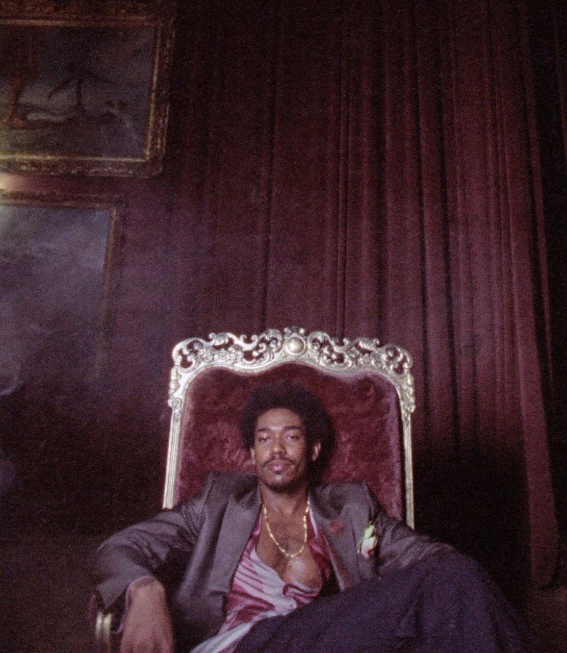 A seated figure in a suit in a tall velvet chair against dark red curtains.