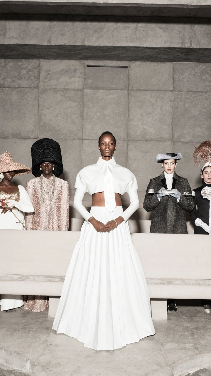 A woman in a sculptural white outfit stands in front of four figures wearing elaborate hats.