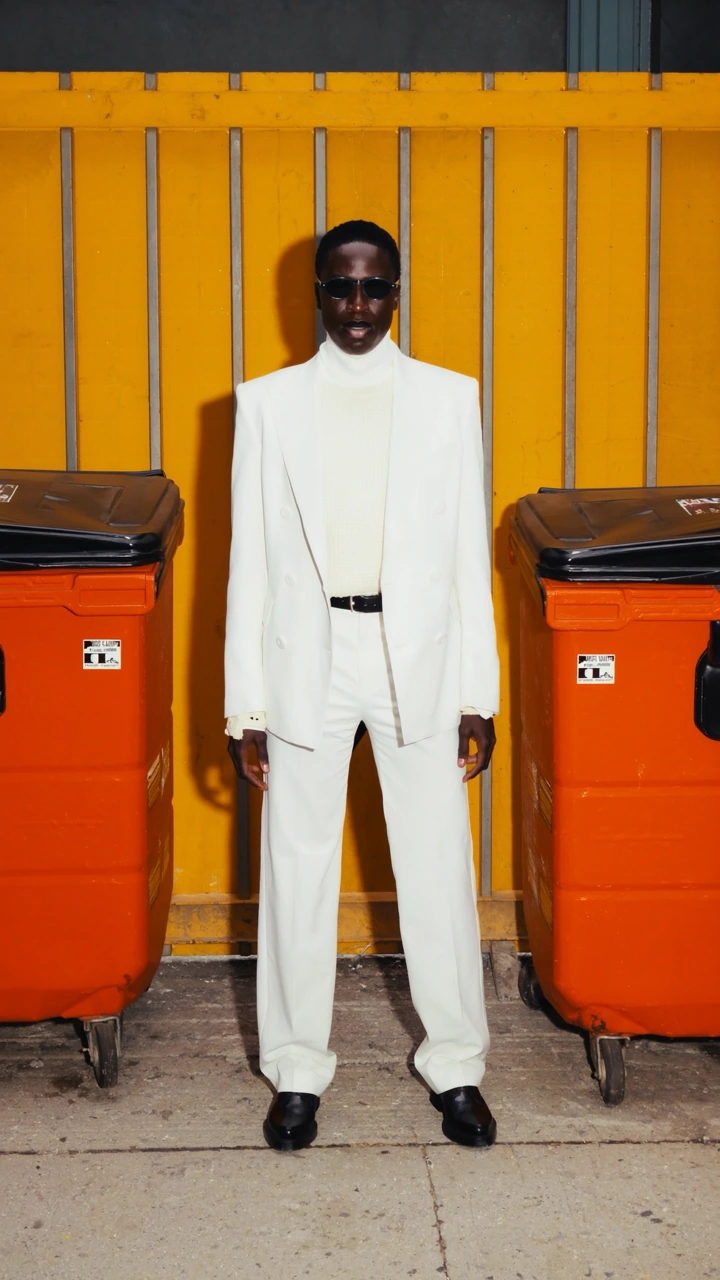 A figure in a white suit and sunglasses between orange bins against a yellow wall.
