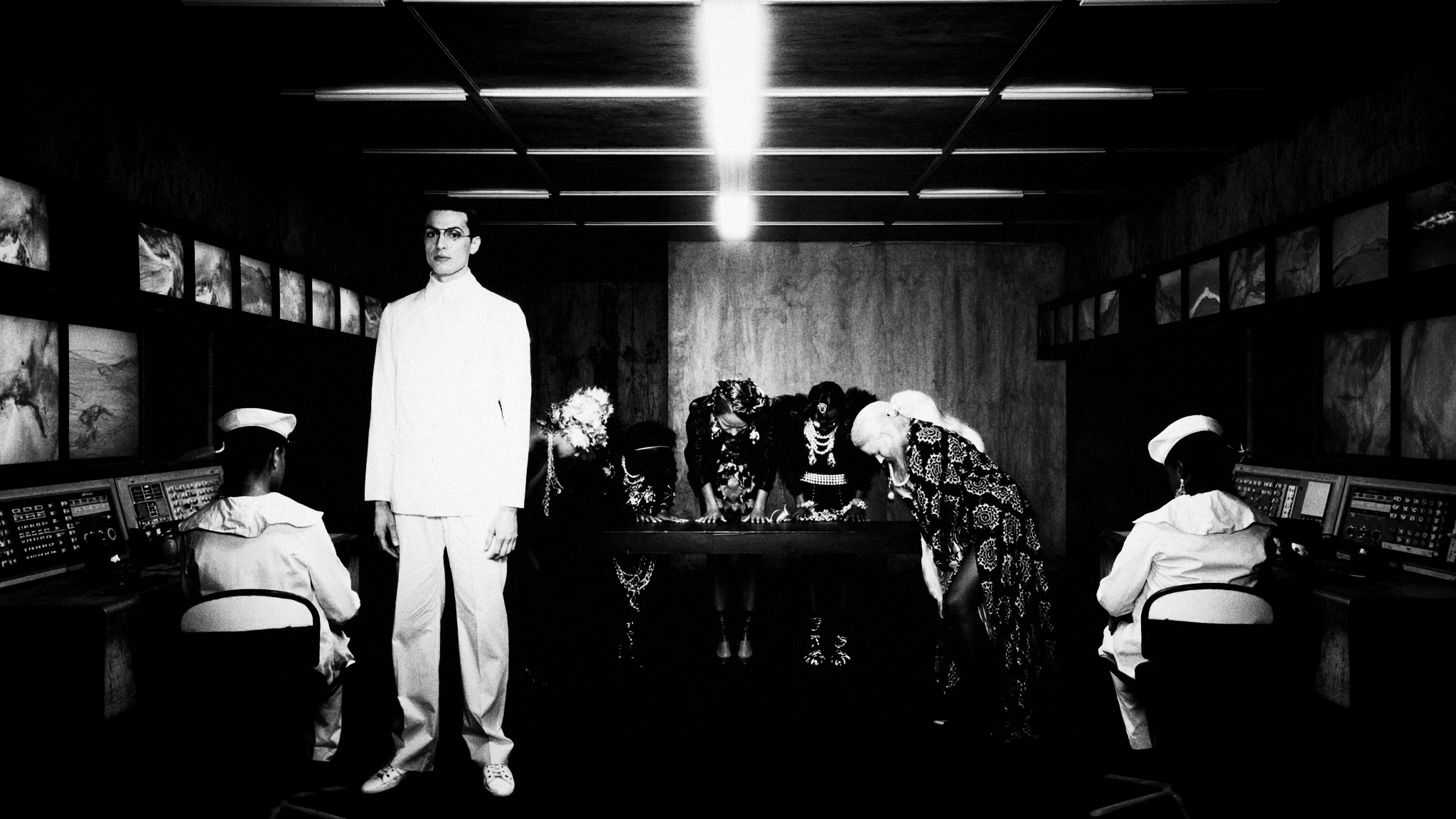 A man in white stands beneath a line of lights in a dark room, surrounded by seated figures.