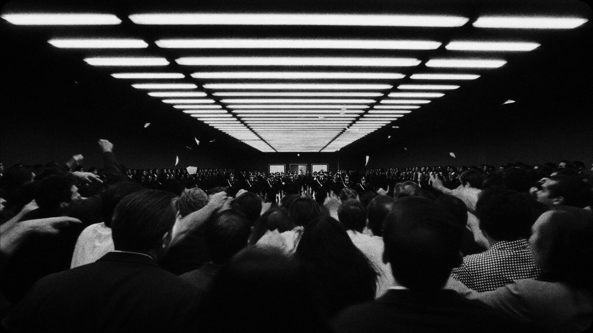 A crowd fills a dark room beneath a luminous ceiling that stretches into the distance.