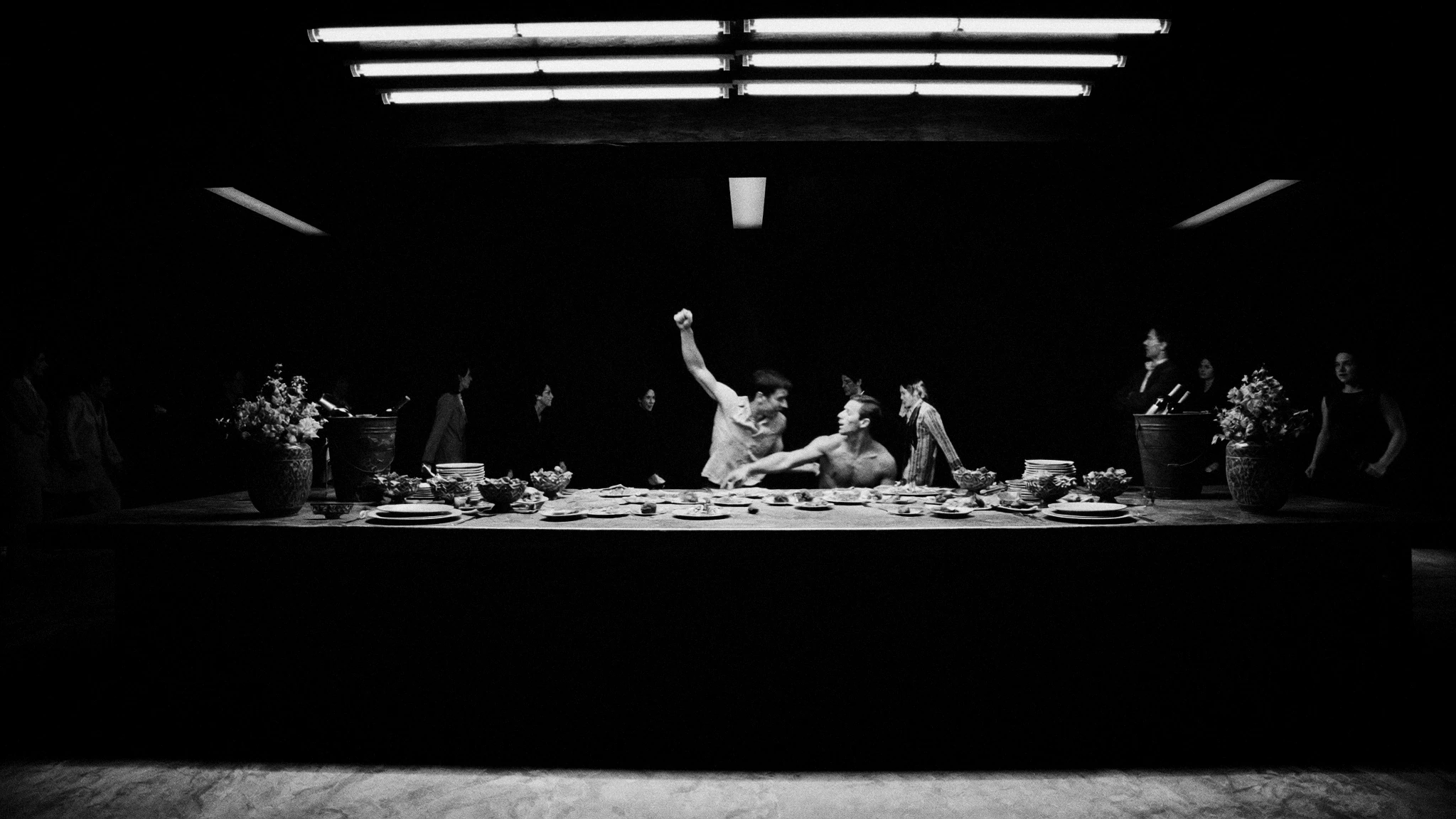 A long table and its seated figures emerge from darkness beneath overhead lights.