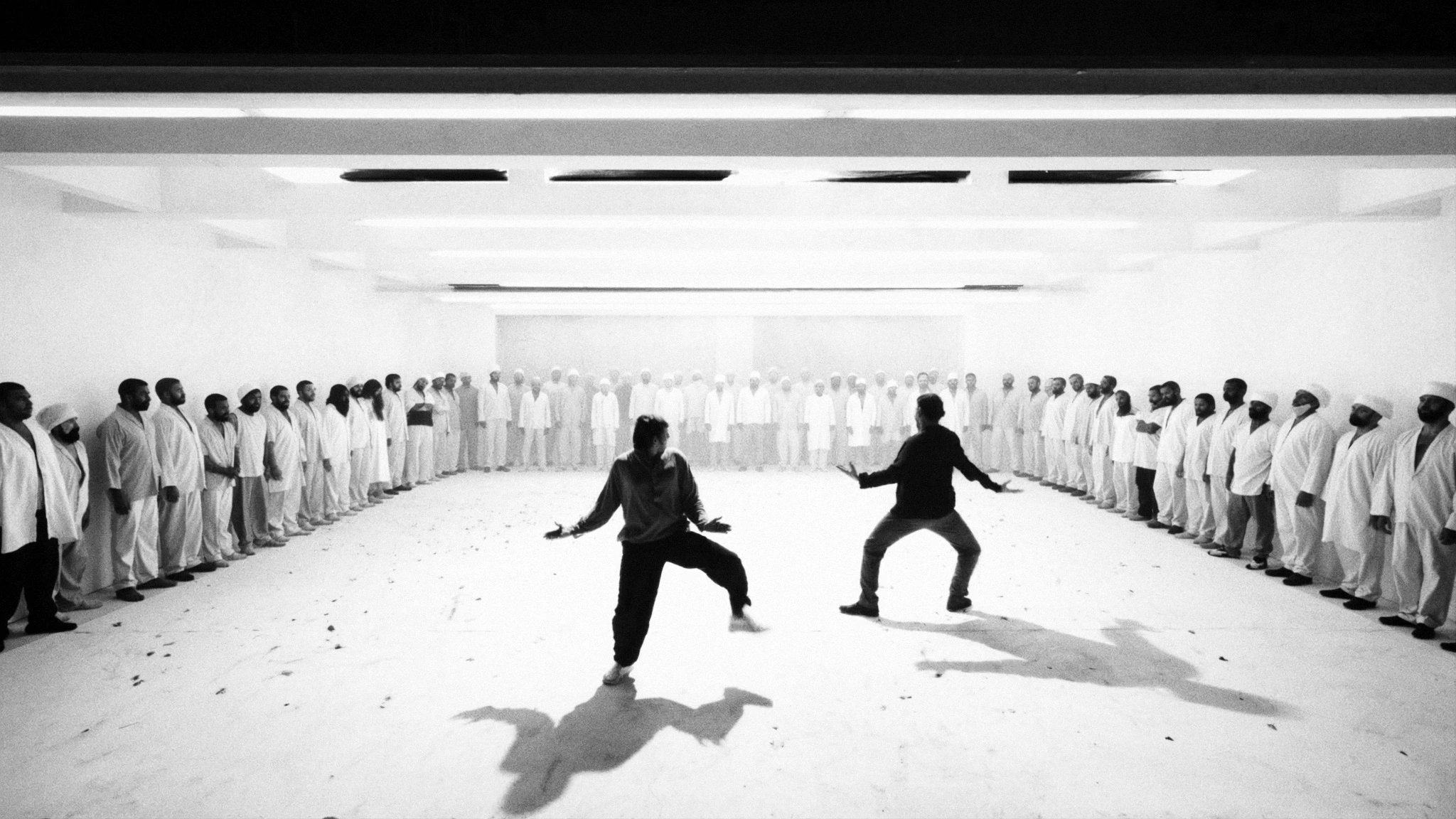 Two figures face one another in a bright room, watched by a surrounding crowd dressed in white.