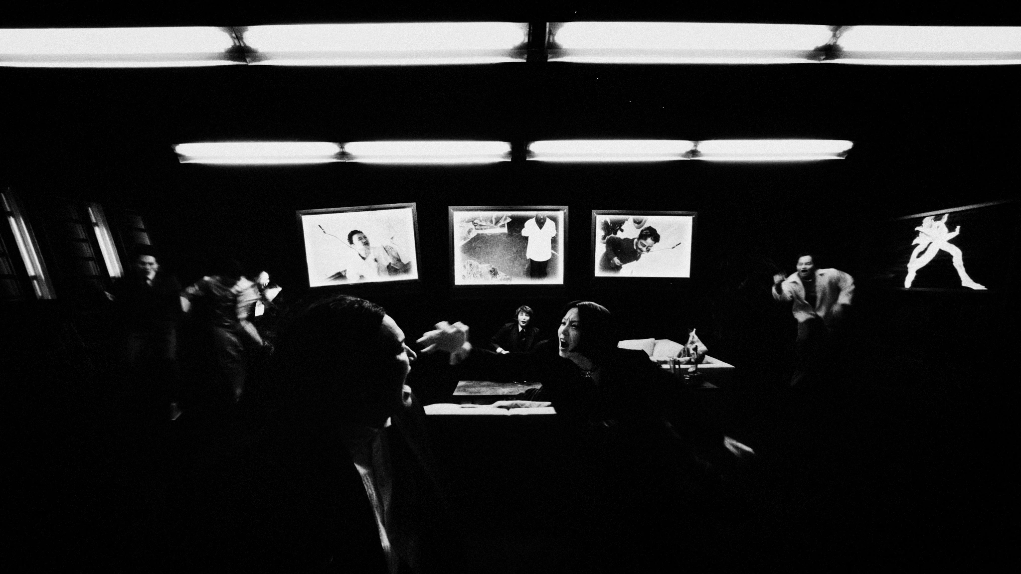 Figures gather beneath lights and suspended picture frames in a dark room.