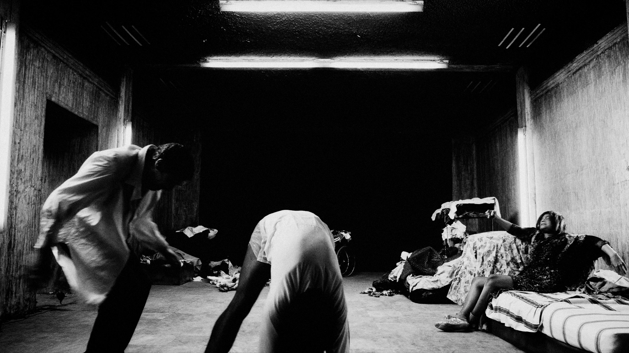 Strange figures sit and move through a rough-walled room under a single fluorescent light.