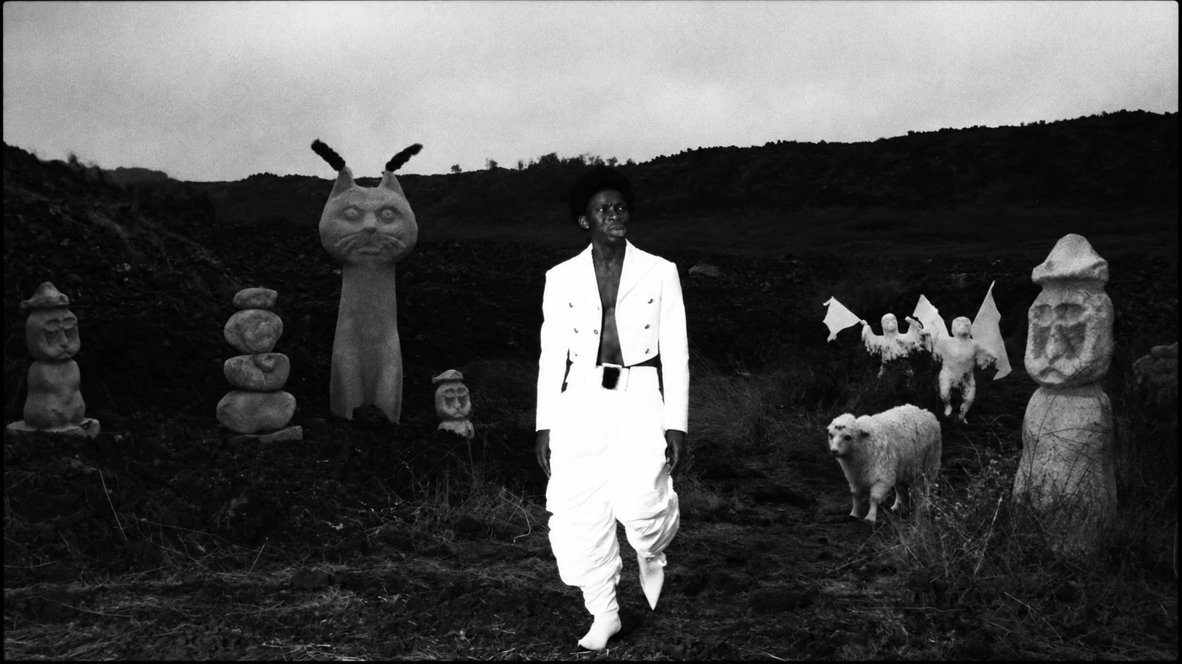 A man in a white suit stands among sculptural creatures on a dark hillside.